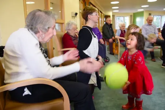 Een inwoonster van het verzorgingstehuis gooit de bal over met een peuter uit de dagopvang.