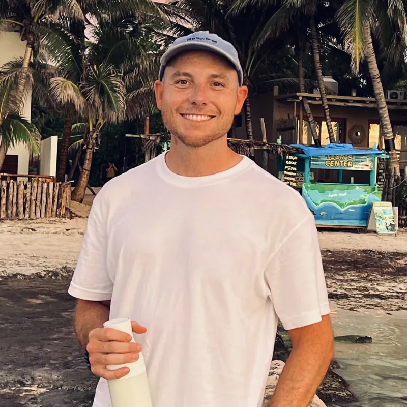 Man met pet kijkt recht in de camera op de achtergrond strand