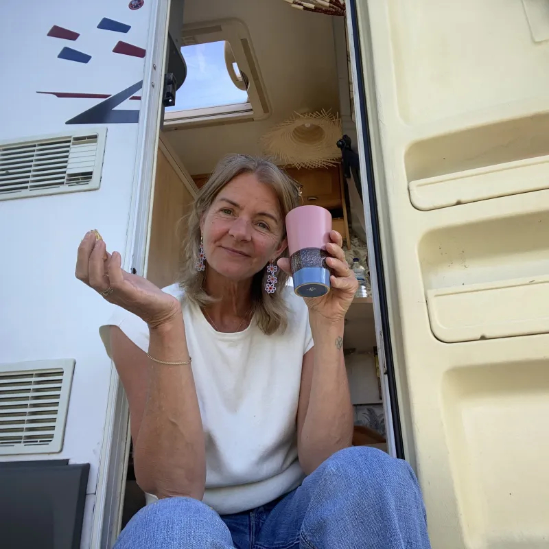 Vrouw zit in de deuropening van een camper en kijkt vriendelijk de camera in
