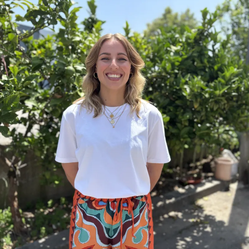 Vrouw in wit shirt voor een heg lachend naar camera