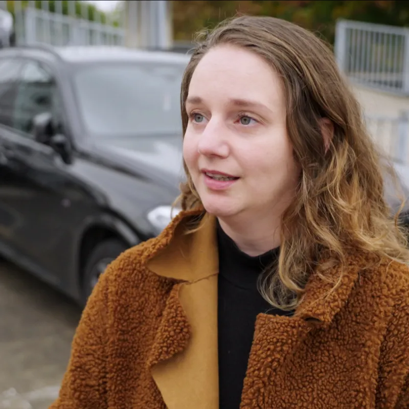 een vrouw in een bruin jasje met een auto op de achtergrond