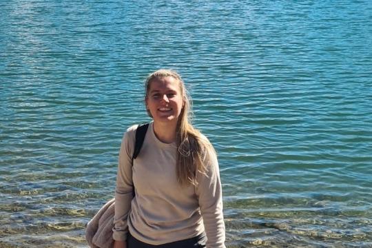 Onderweg naar liefde - Emma Vrouw staat met hond bij het water en kijkt lachend de camera in