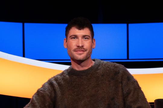 Man met bruin haar en snor en bruine trui