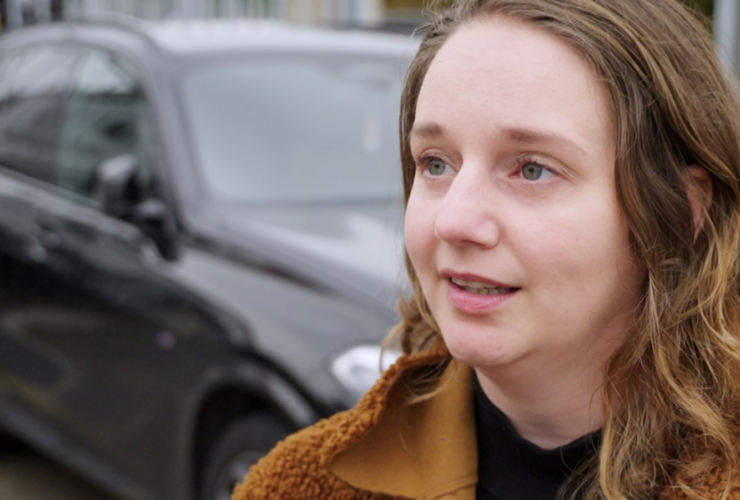 een vrouw in een bruin jasje met een auto op de achtergrond