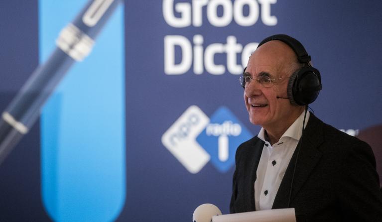 Presentator Frits Spits met microfoon in de hand voor een banner met hierop de tekst Het Groot Dictee