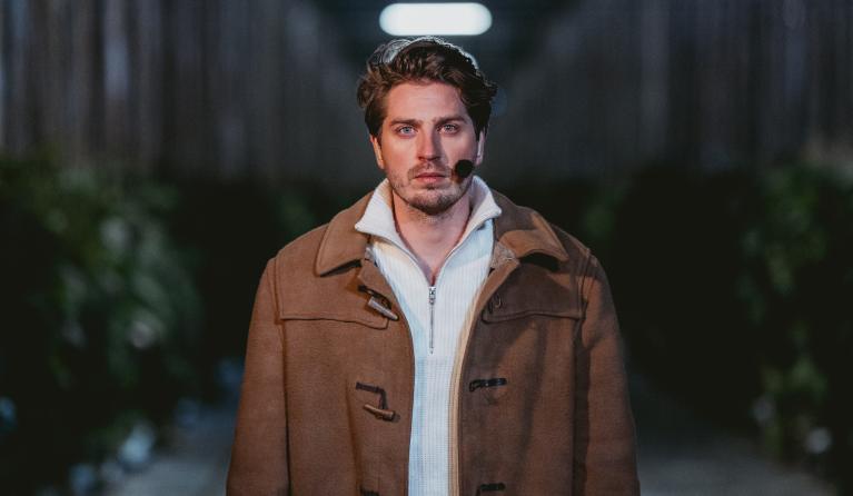 Een man staat op een afstandje serieus de camera in te kijken