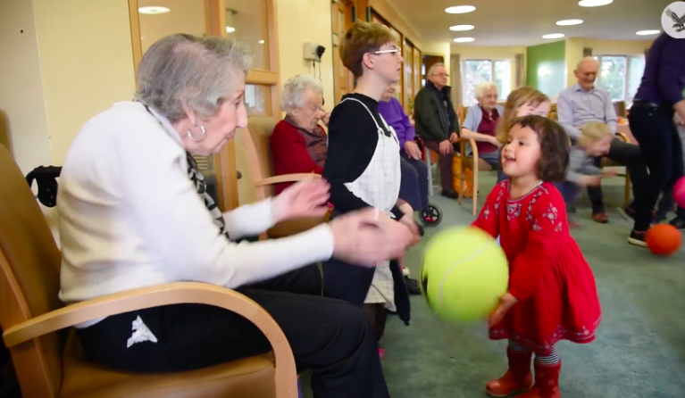 Een inwoonster van het verzorgingstehuis gooit de bal over met een peuter uit de dagopvang.