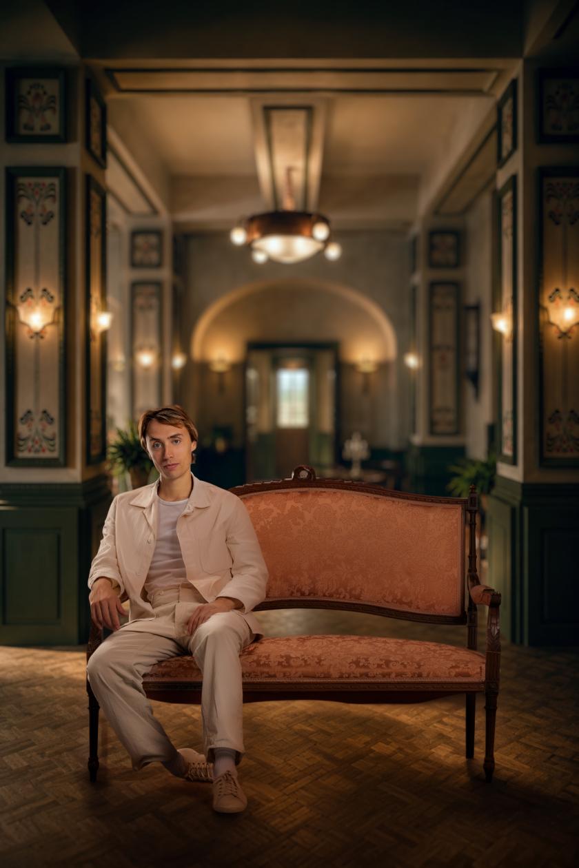 Een man in beige pak poseert op een bank in een luxe hotel