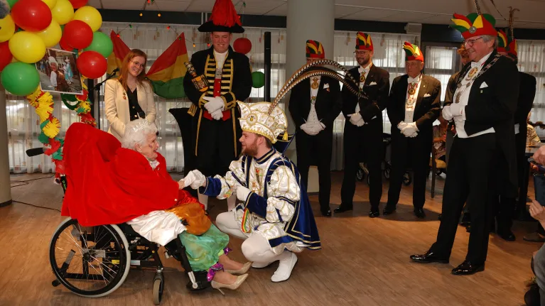 Mevrouw in rolstoel omringd door mannen in carnavalspakken