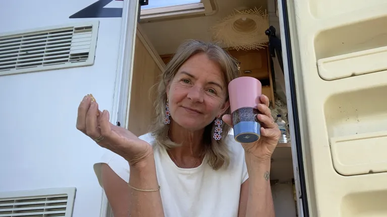 Vrouw zit in de deuropening van een camper en kijkt vriendelijk de camera in
