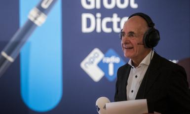 Presentator Frits Spits met microfoon in de hand voor een banner met hierop de tekst Het Groot Dictee