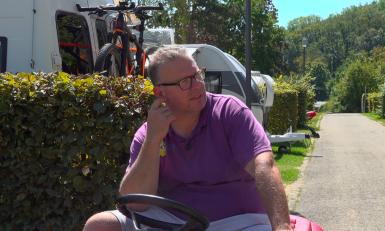 Een man met paars shirt op een grasmaaier op een camping