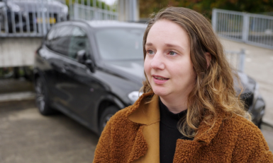 een vrouw in een bruin jasje met een auto op de achtergrond